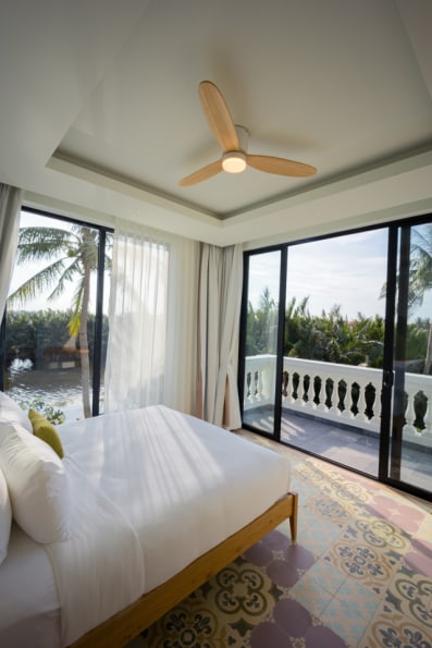 A bright and airy bedroom features large glass doors opening to a private balcony, offering views of tropical greenery. A ceiling fan is positioned above the spacious bed, which is complemented by textured bedding. Colorful tiles add a touch of charm to the floor.