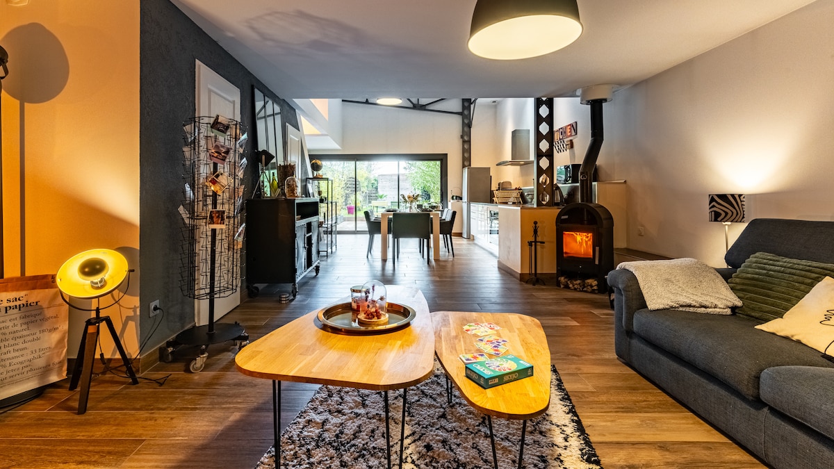 A modern living area features wooden coffee tables and a cozy seating arrangement. A wood stove adds warmth, while large windows provide a view of the garden. The open layout connects to the kitchen and dining space, enhancing the bright atmosphere.