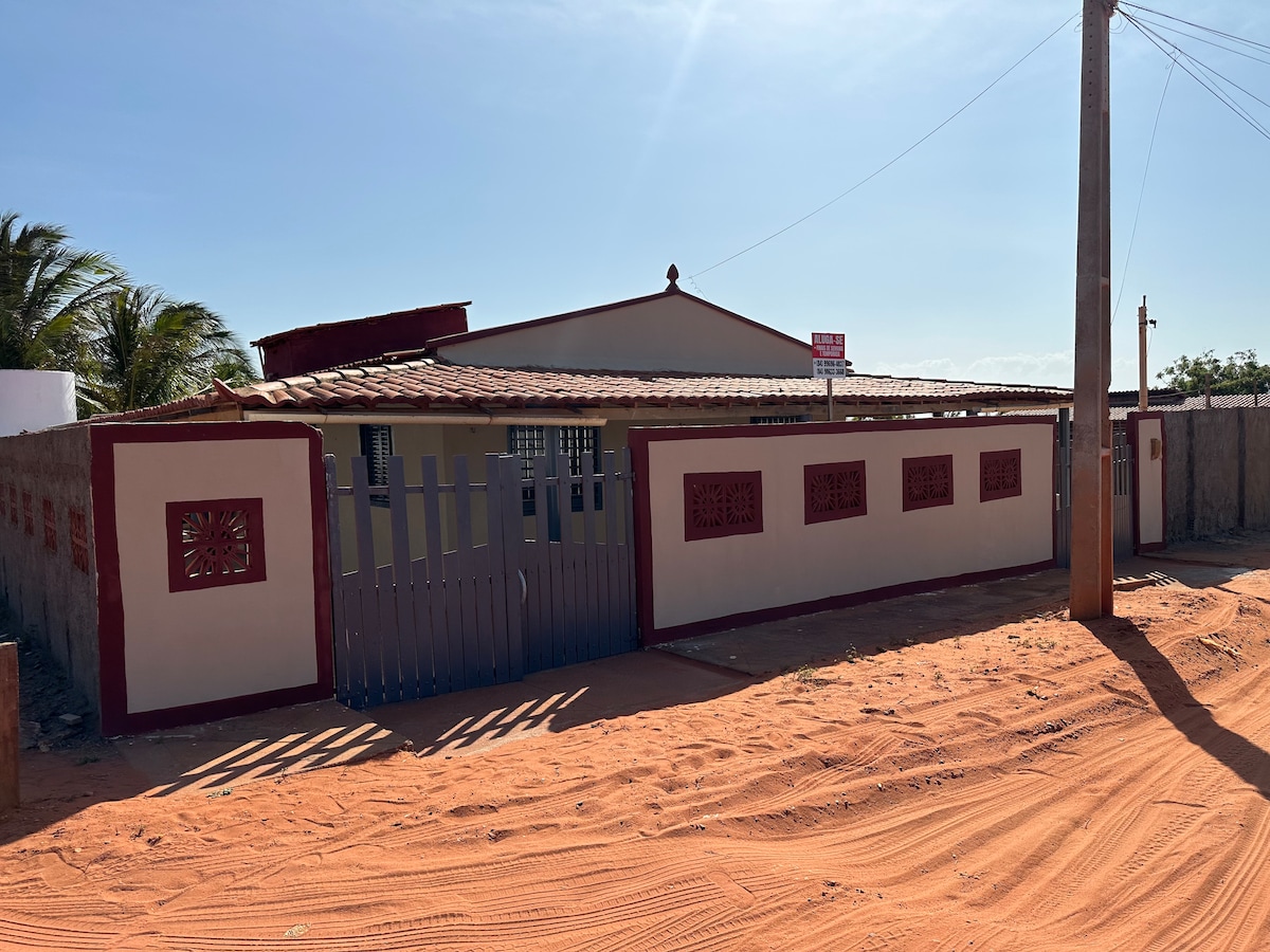 A welcoming exterior view of the property is shown, featuring a home with a red-tiled roof and decorative wall elements. The gated entrance offers access to a sandy pathway leading to the house, surrounded by greenery under a clear blue sky.
