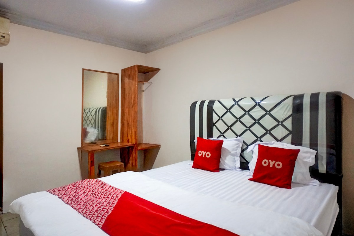 A modern bedroom features a spacious bed with white linens and decorative red accent pillows. A wooden dresser with a mirror is positioned against the wall, providing a functional space for guests. Natural light is filtered through a nearby window, enhancing the clean atmosphere.