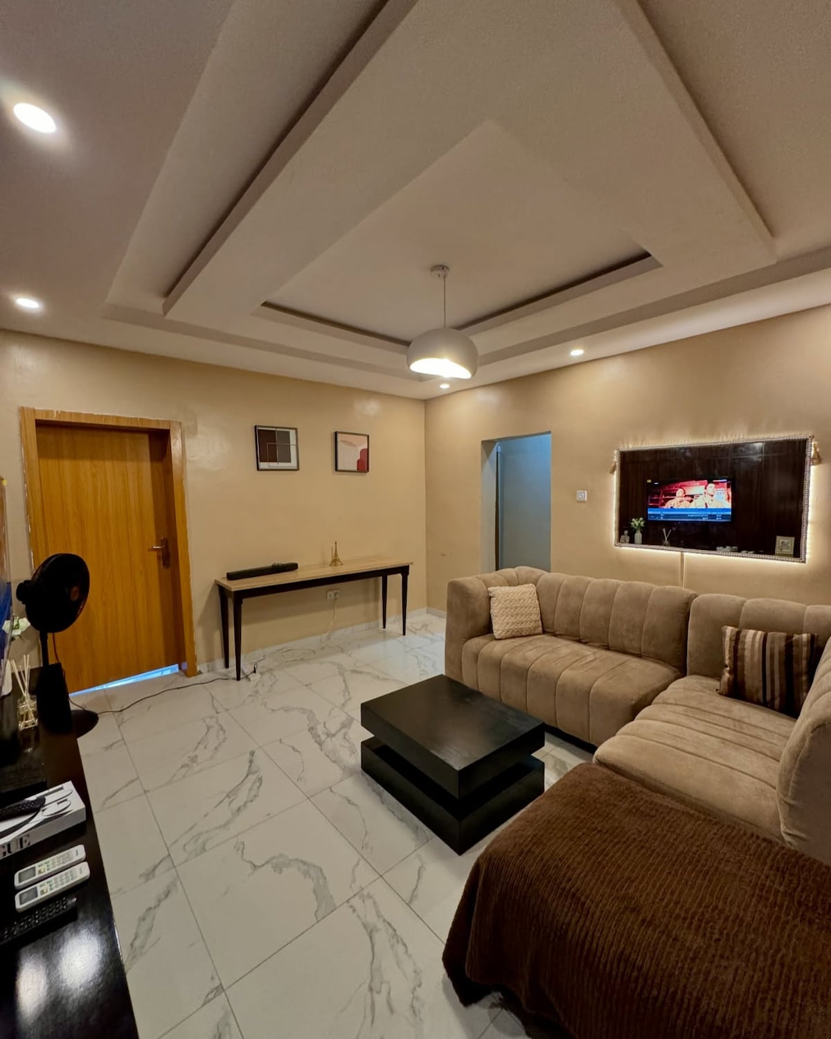 A modern living area features a spacious sectional sofa in neutral tones, complemented by a sleek black coffee table. A flat-screen TV is mounted on the wall, and the room is illuminated by a contemporary ceiling light. Warm wooden accents are visible in the open doorway.