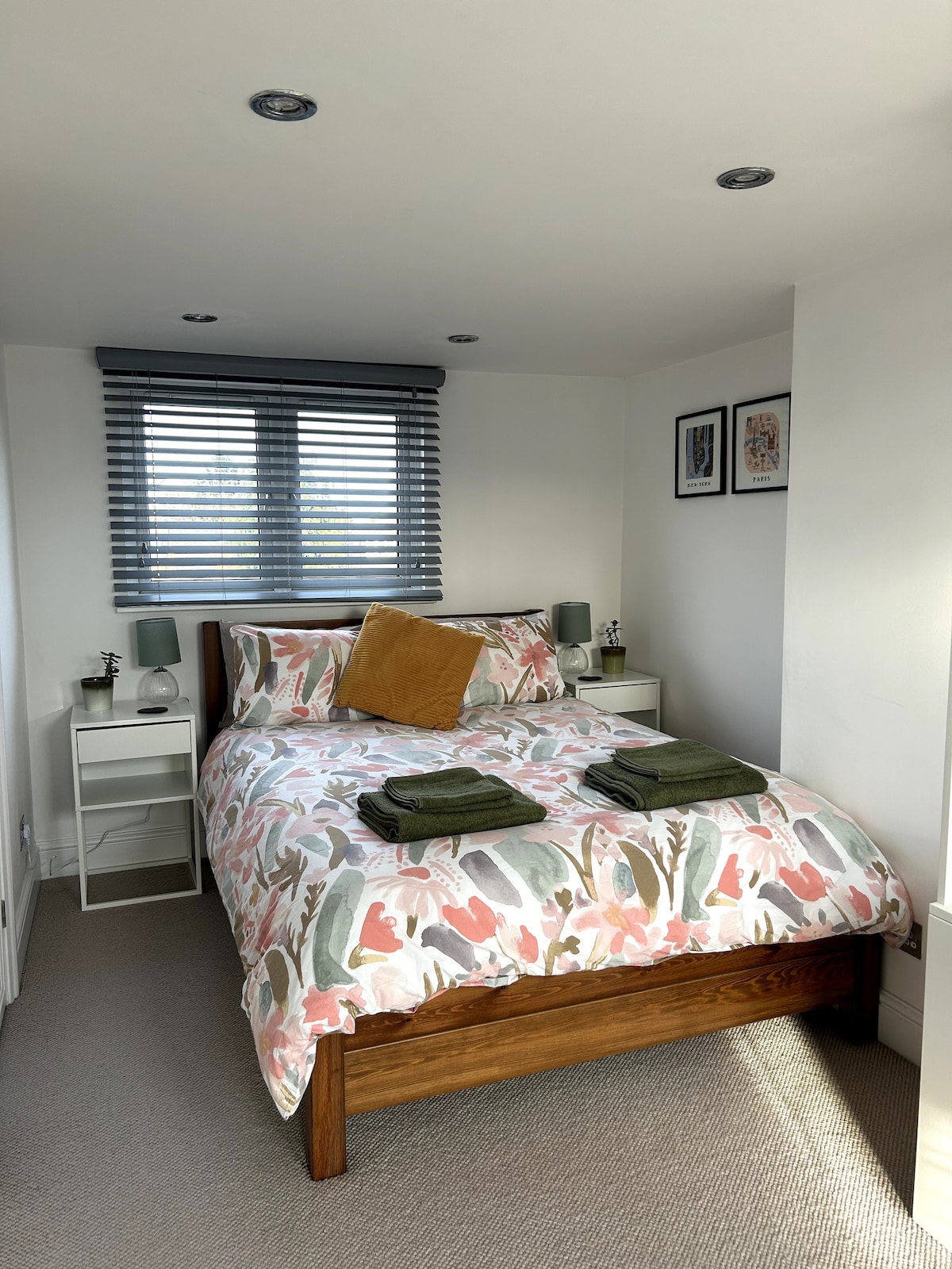 A cozy loft room features a wooden bed with a floral duvet cover, accented by throw pillows. Two nightstands hold lamps, while framed artwork adorns the wall. Natural light enters through a window adorned with blinds, creating a serene atmosphere.