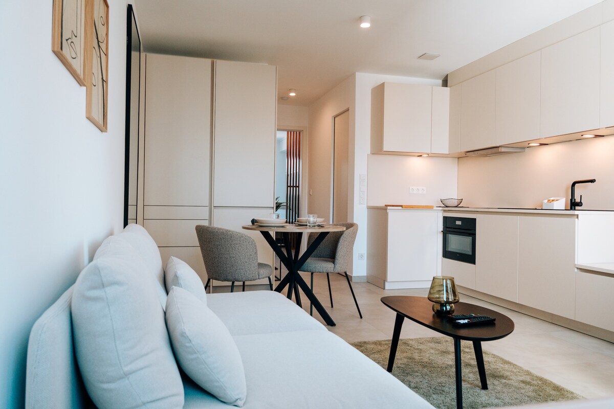 A modern living space features a light-colored sofa, a round dining table with two chairs, and a well-equipped kitchen area. Neutral tones are complemented by soft lighting. The design prioritizes functionality and comfort, creating an inviting atmosphere for relaxation or meals.