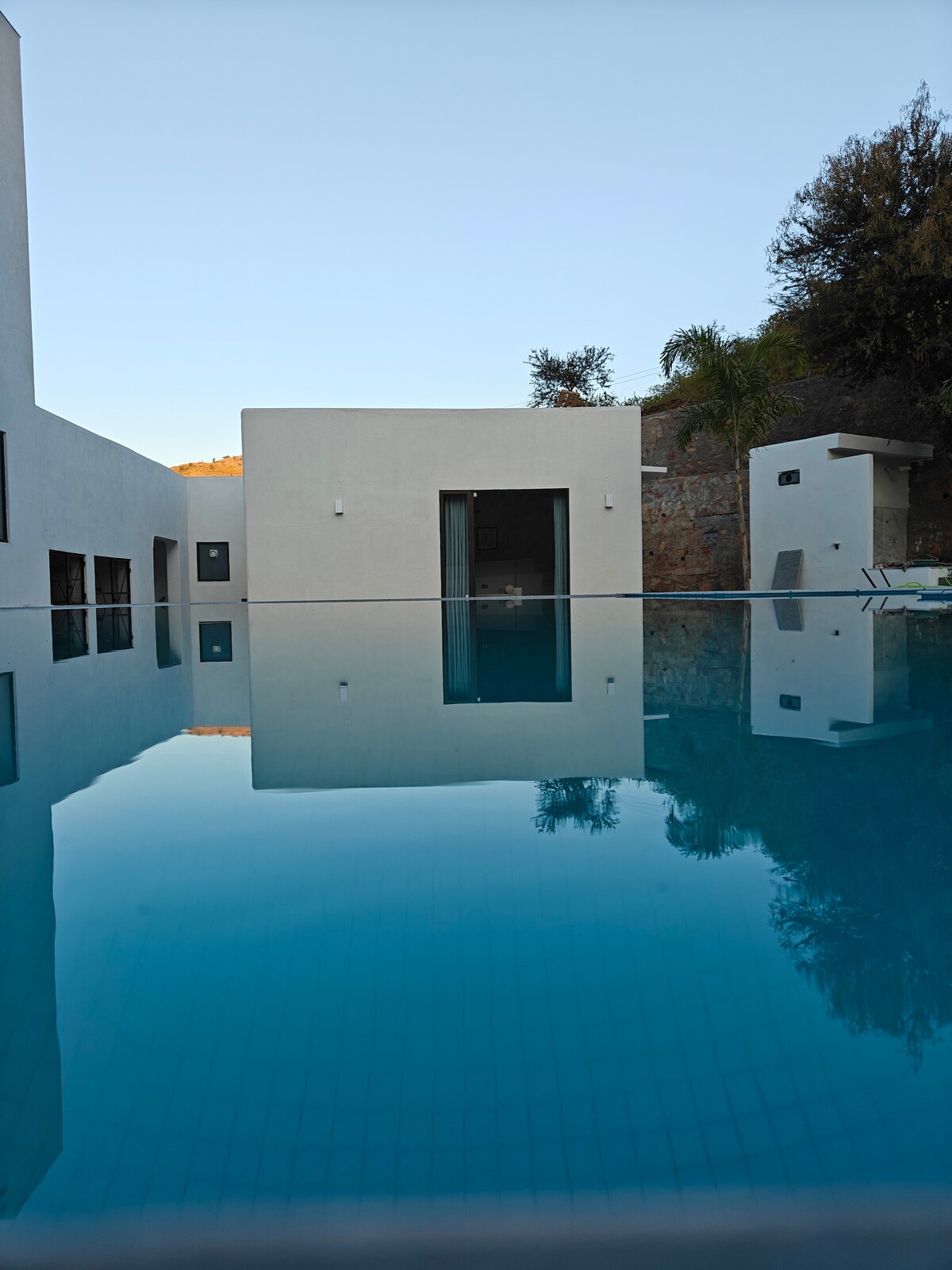The image showcases a serene swimming pool reflecting the surrounding architecture and greenery. The water appears calm and crystal clear, enhancing the tranquil atmosphere. A modern building with large glass doors is visible in the background, complemented by natural elements on the property.
