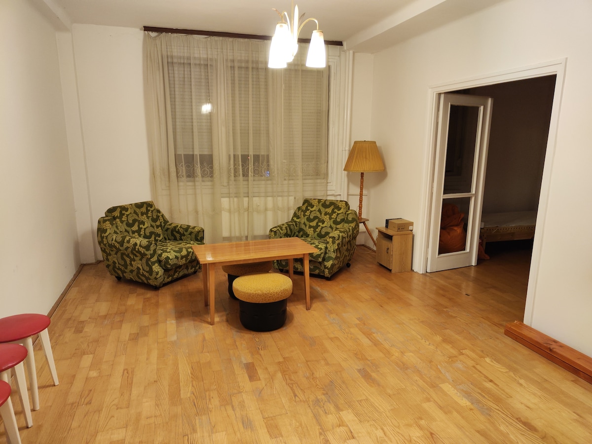 A spacious living area is furnished with two patterned bean bag chairs and a wooden coffee table. Natural light filters through sheer curtains, illuminating the light wooden floor. A floor lamp stands against the wall, and a doorway leads to another room.
