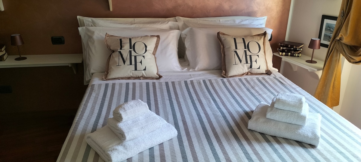 A neatly made bed is presented with white linens and decorative throw pillows featuring the word 'HOME.' Soft, folded towels are placed at the foot of the bed. A warm, neutral wall color complements the overall aesthetic, and bedside lamps can be seen on either side.