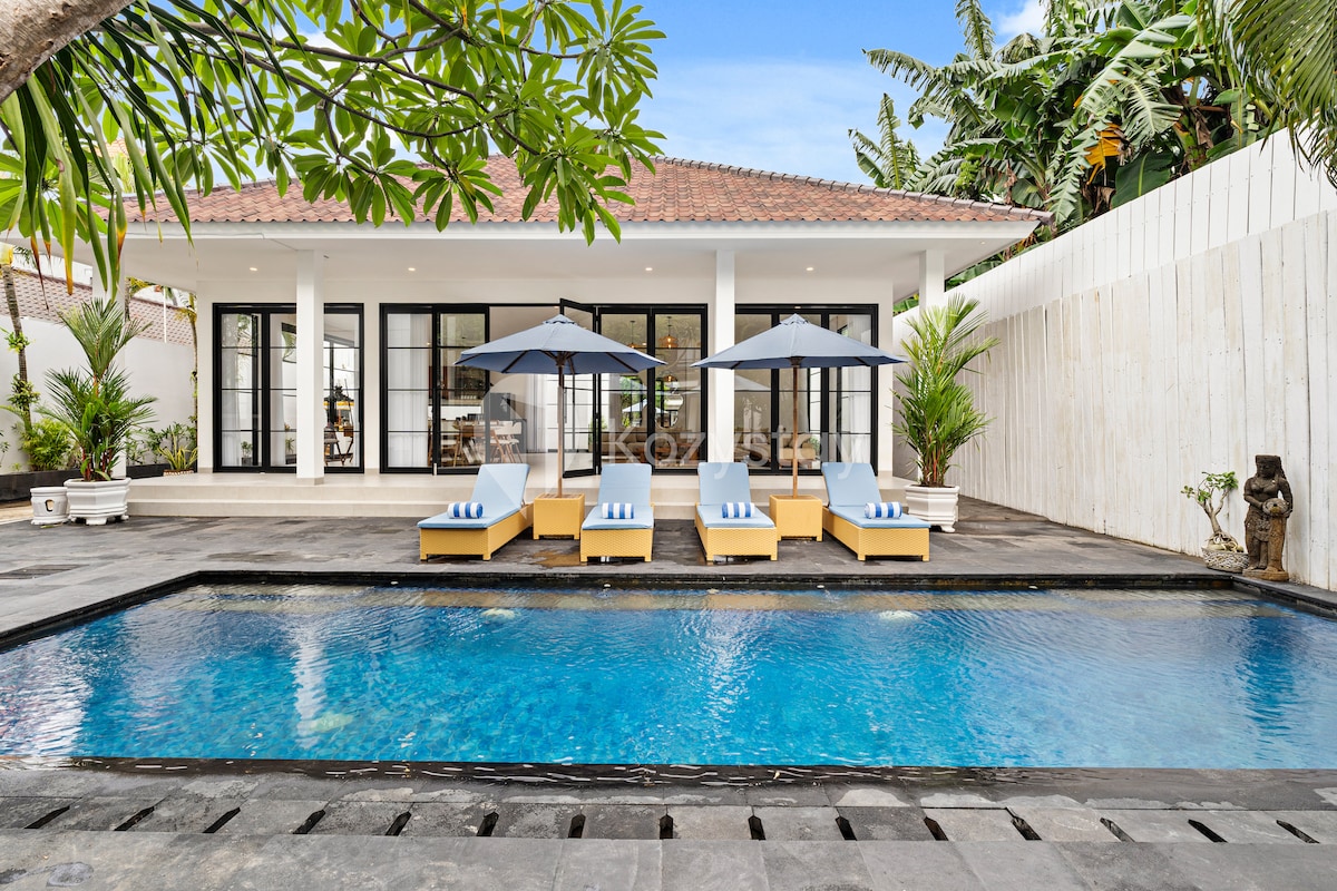 A private outdoor swimming pool with sun loungers is surrounded by tropical greenery. Two large umbrellas provide shade over the loungers. The villa's glass doors create a seamless connection to the indoor living spaces, enhancing the bright and open atmosphere.