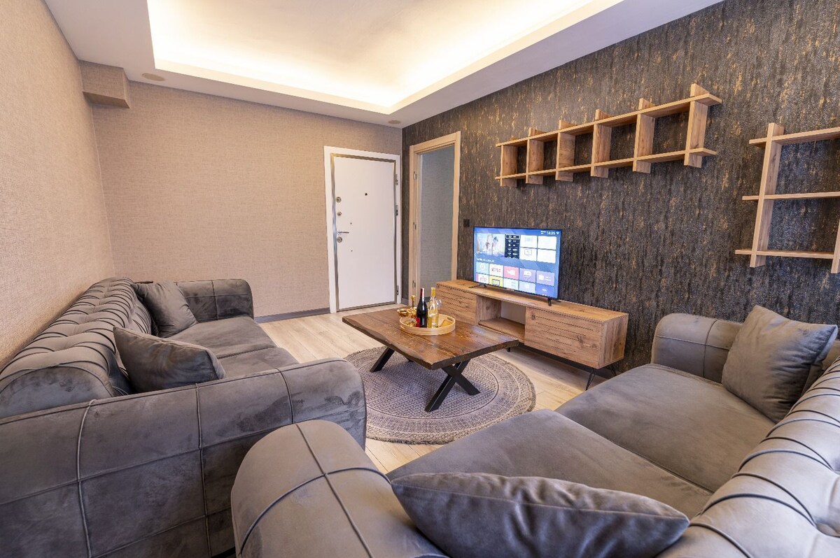 A spacious living area features two large sofas arranged around a central coffee table. A flat-screen TV is mounted on the wall, with wooden shelving units providing storage. Light from overhead lighting creates a comfortable ambiance in the neutral-toned room.