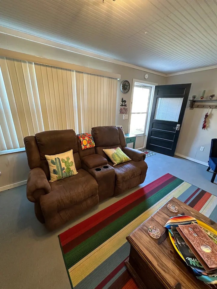 A cozy living area features two brown recliners adorned with decorative cushions. A colorful area rug lies beneath a wooden coffee table, enhancing the space's warmth. Large windows with vertical blinds allow natural light, while a dark door provides access to the outside.