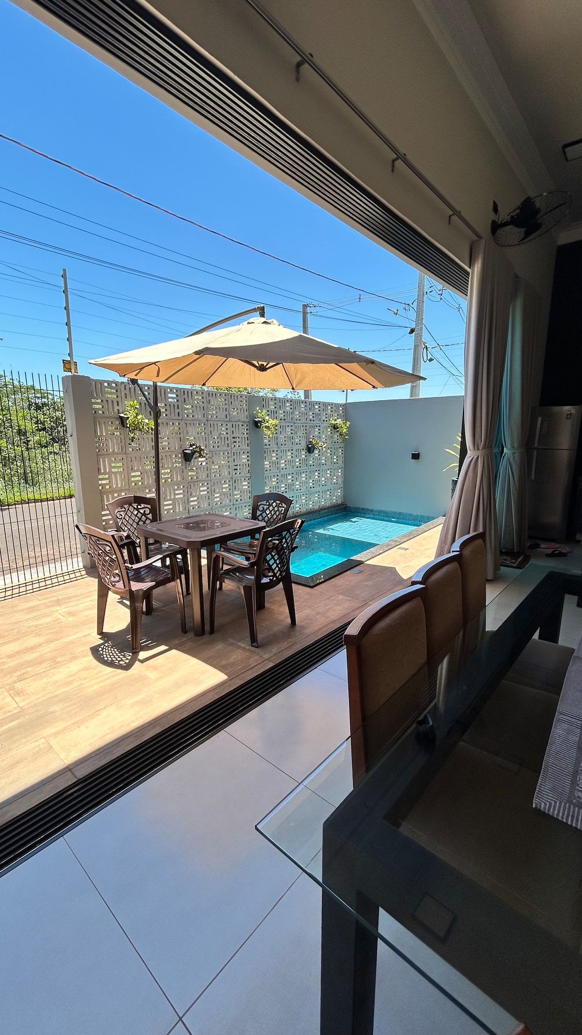 A dining area features a glass table with four chairs, positioned adjacent to large open doors leading to a private patio. The outdoor space includes a small pool, an umbrella, and a decorative privacy screen, framed by clear blue skies.