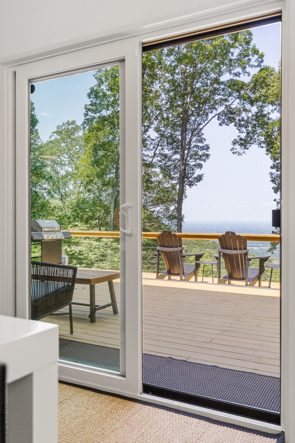 Sliding glass doors open to a spacious deck overlooking a scenic view. Comfortable outdoor seating is visible, framed by lush trees that enhance the natural setting. A grill is positioned on the deck, providing options for outdoor dining.