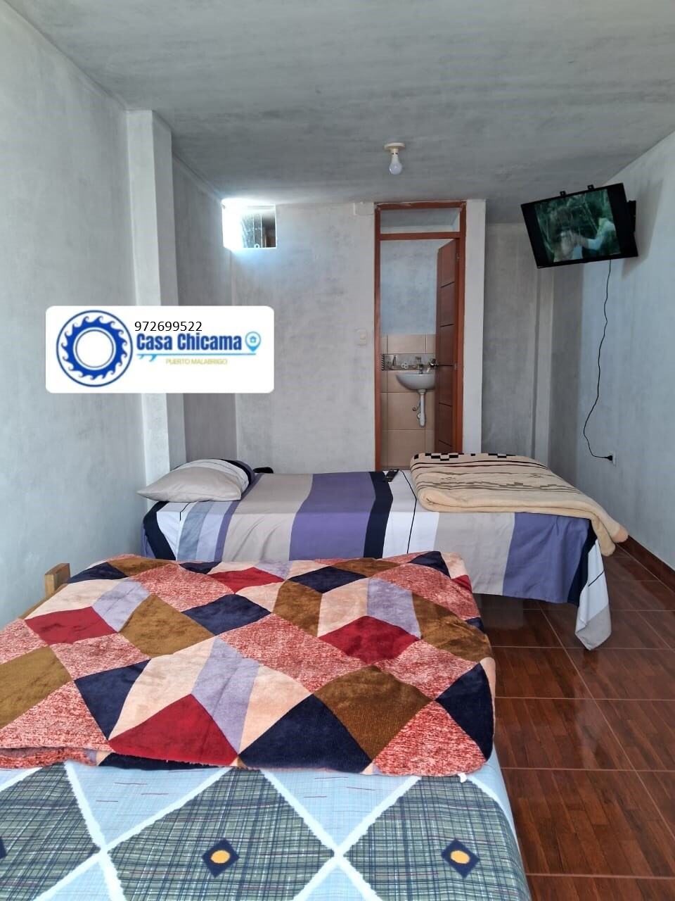 A simple sleeping area features two twin beds covered with colorful blankets. A television is mounted on the wall, while a small bathroom is visible through an open door. Neutral-colored walls and wooden flooring create a calm environment.