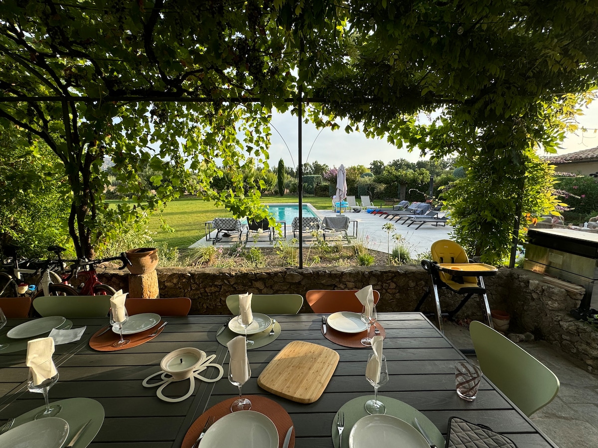 An outdoor dining area features a large table set with plates and napkins, surrounded by greenery. The backdrop reveals a heated pool with sun loungers and a play area, all under the shade of a vine-covered trellis.