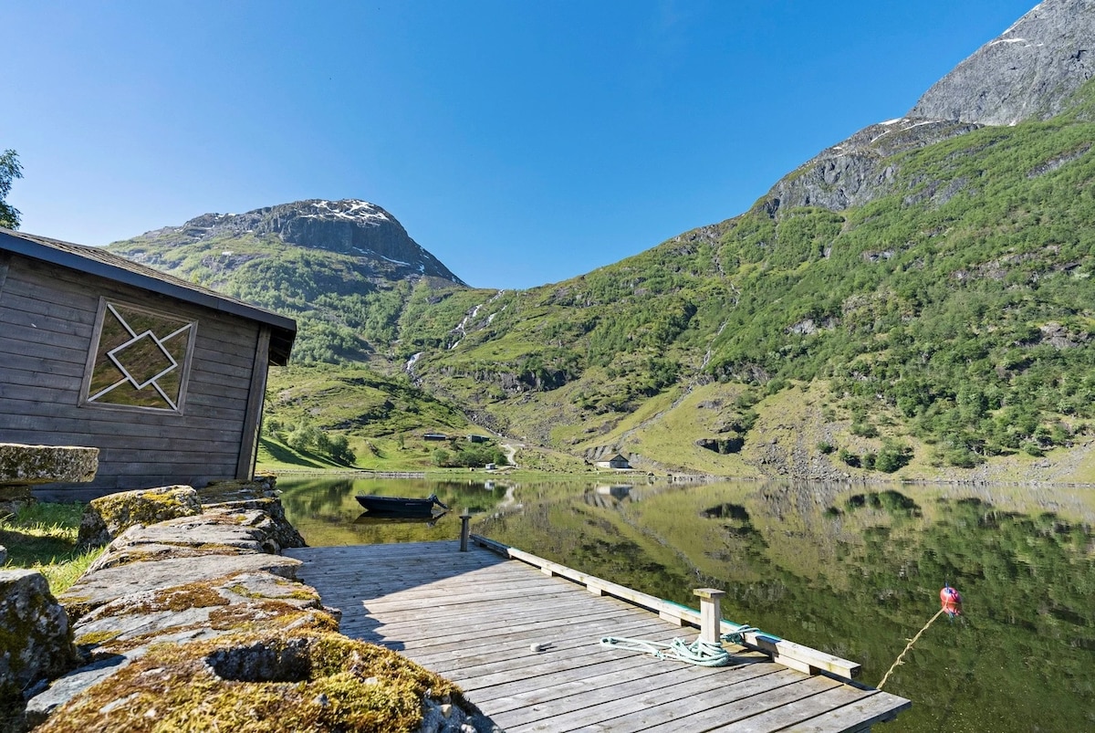 A wooden deck extends over still water, reflecting the surrounding mountains and lush greenery. On the left, a cozy building characterized by a geometric emblem is visible. A small boat rests nearby, contributing to the serene ambiance of the natural landscape.