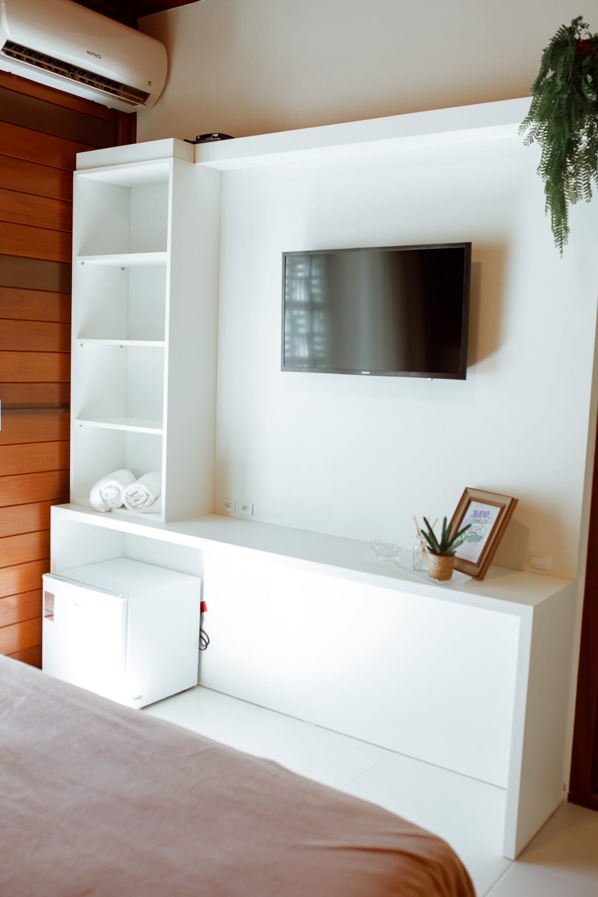 A modern entertainment unit is displayed, featuring a flat-screen TV mounted on a white wall. Below, a compact refrigerator is visible alongside neatly arranged towels. A small potted plant and a framed picture add subtle decor, complemented by warm wooden accents in the background.