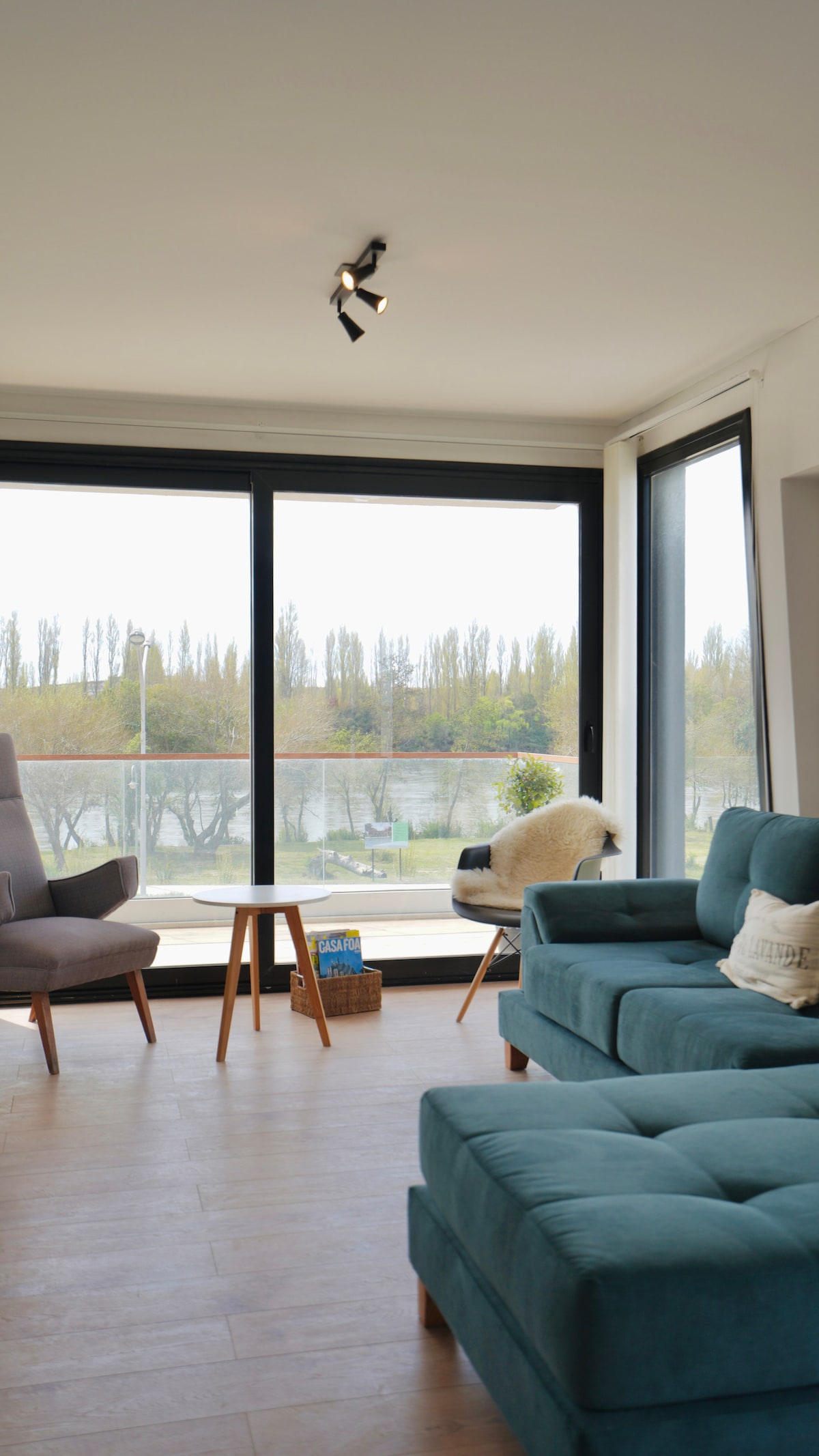 A modern living area is shown, featuring a large window that allows natural light to fill the space. Soft blue seating is arranged comfortably, complemented by a small wooden table. Green trees can be seen outside, adding a touch of nature to the scene.