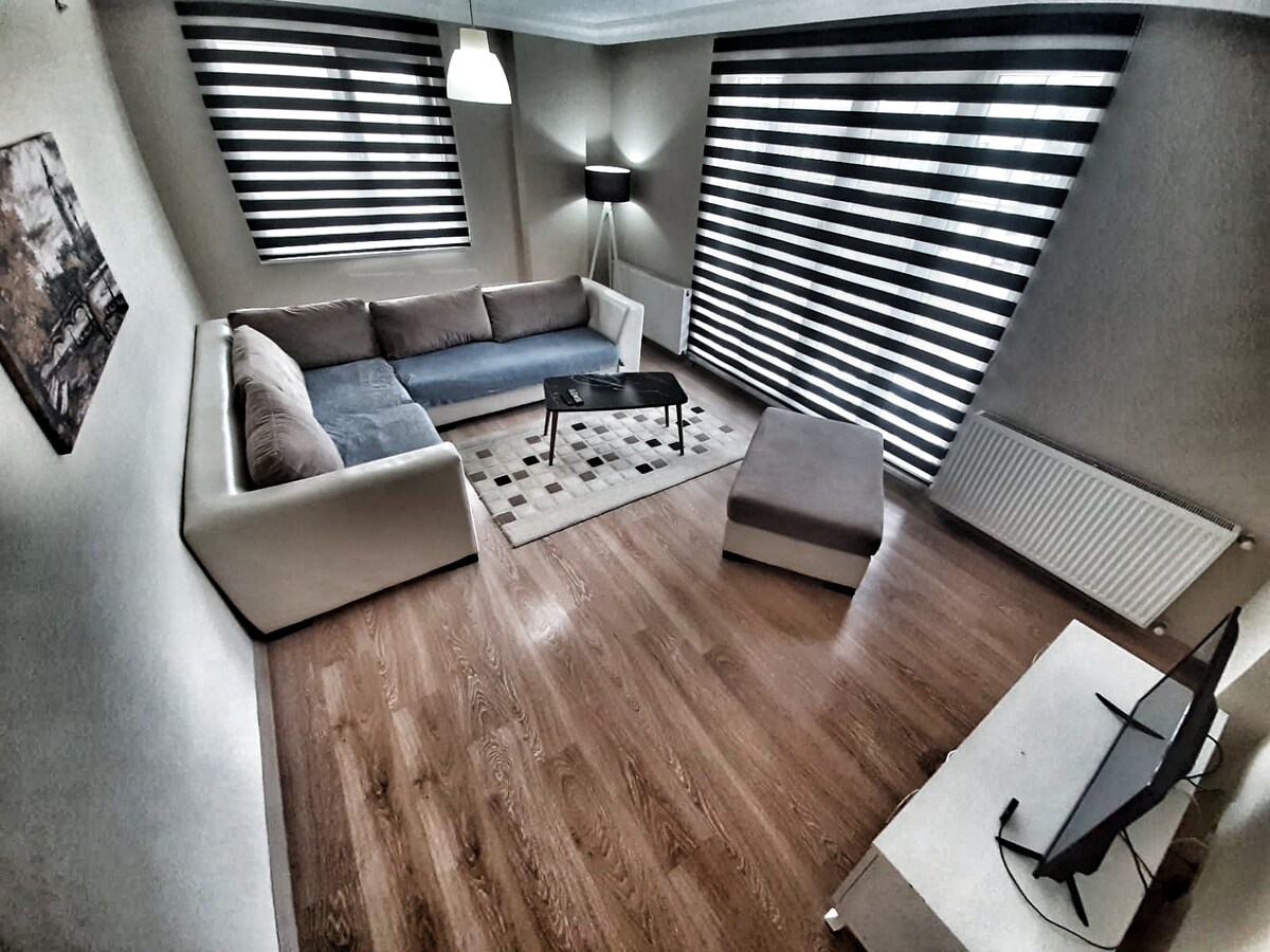 A modern living area features a sectional sofa in muted tones, complemented by a simple coffee table on a patterned rug. Large windows with striped blinds allow natural light to fill the room, while a standing lamp adds a soft glow to the space.