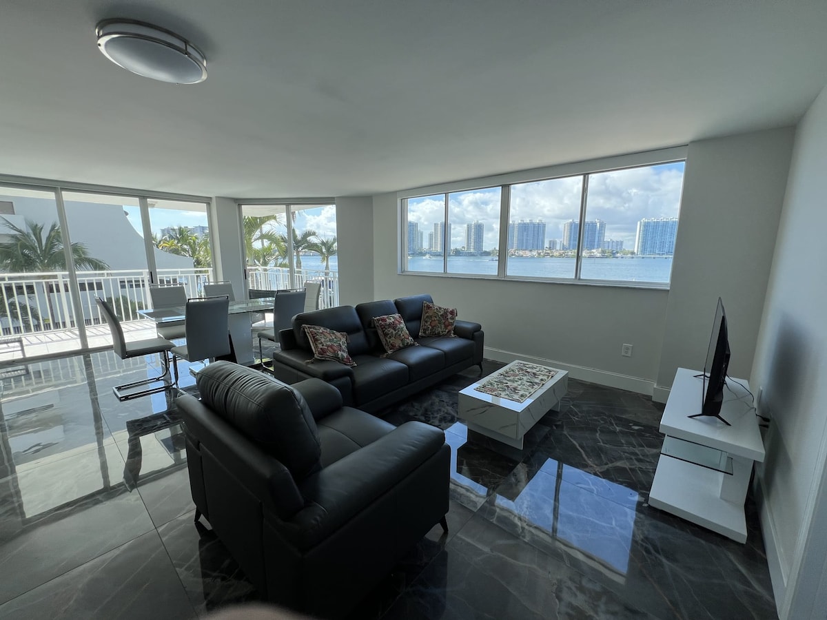 A spacious living room features large windows offering views of the bay. Two black sofas are arranged around a coffee table, while a TV sits on a white stand. The marble flooring adds a touch of elegance, and furnishings are complemented by a light color scheme.