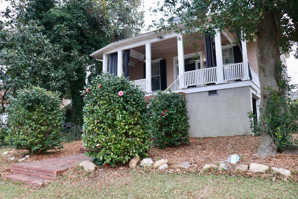 A charming historic home is presented with a welcoming porch featuring white columns and a combination of open and draped windows. Lush green bushes frame the staircase leading to the entrance, while a path made of bricks leads to the house, surrounded by a natural landscape.