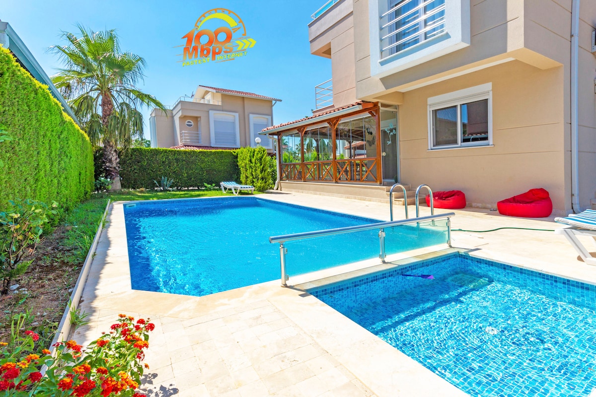 A private pool is surrounded by beautifully manicured landscaping, featuring a mix of vibrant flowers and lush green hedges. Comfortable bean bags are positioned near the pool for relaxation, while a covered outdoor seating area provides a shaded spot for lounging.
