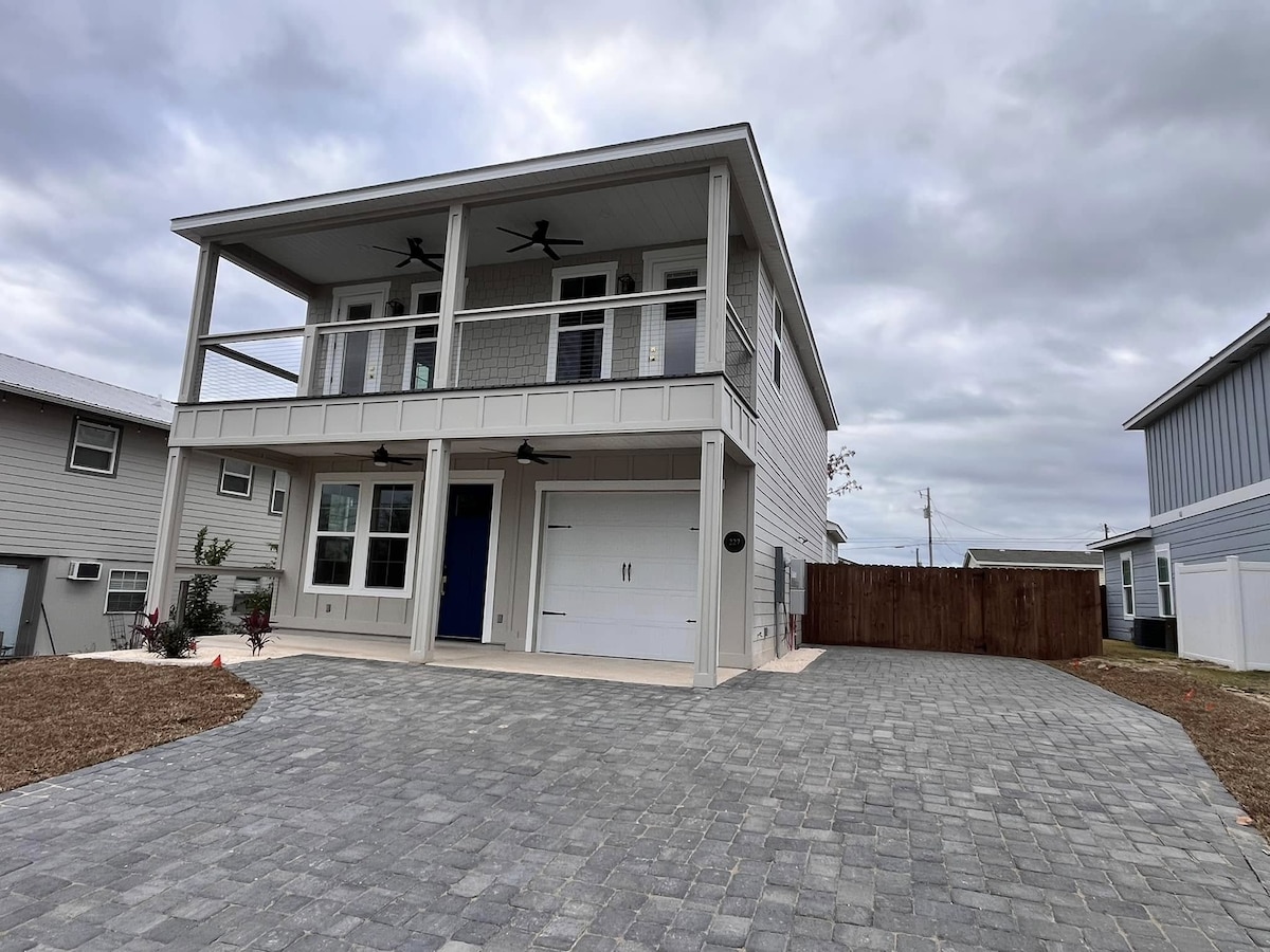 A two-story house showcases a modern design with a light grey exterior. A covered front porch is supported by columns, with ceiling fans visible above. A paved driveway leads to a garage door, while landscaping includes minimal greenery and a wooden fence enclosing the yard.