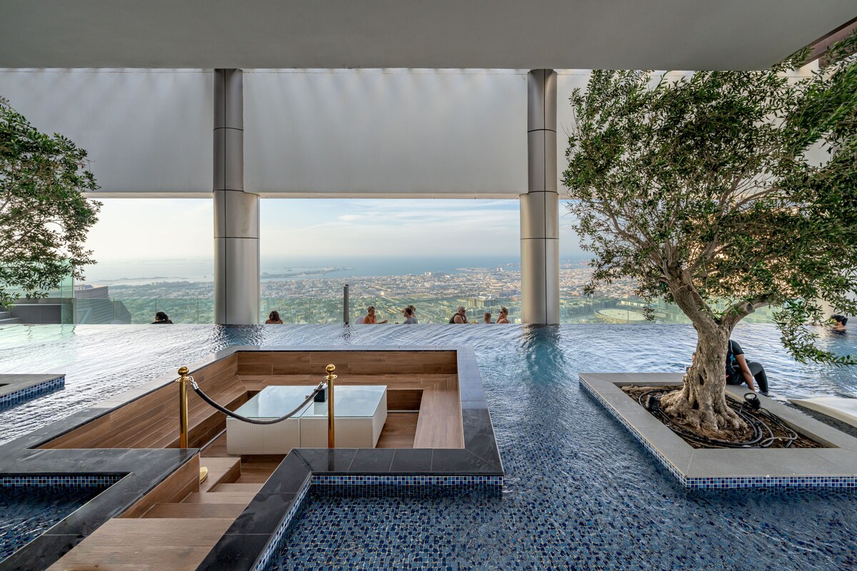A rooftop swimming pool is surrounded by large windows offering panoramic views of the city. Modern wood decking encircles a sunken seating area, while two olive trees are placed in stone planters, adding greenery to the tranquil space.