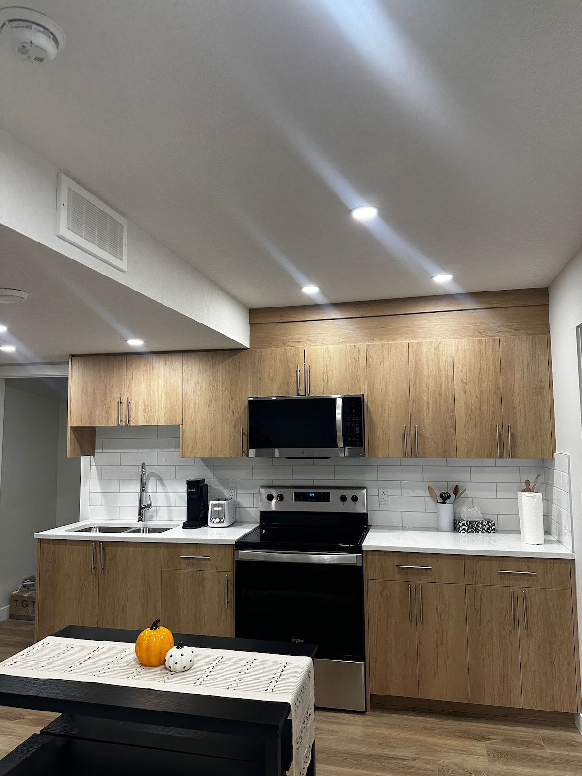 A modern kitchen features light wood cabinetry and sleek white countertops. Stainless steel appliances, including a stove and microwave, complement the design. Natural light is provided by overhead LED lights, creating a bright workspace. Decorative items are arranged on the counter, enhancing functionality.