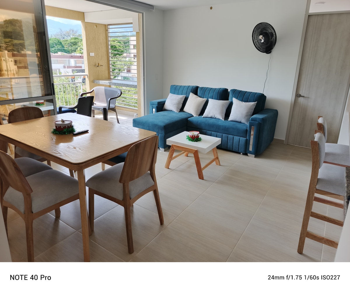 An open living space features a light wood dining table surrounded by four chairs, with vibrant blue sofas providing comfortable seating. Natural light fills the area through large glass doors, leading to a balcony that offers a view of the surrounding landscape.