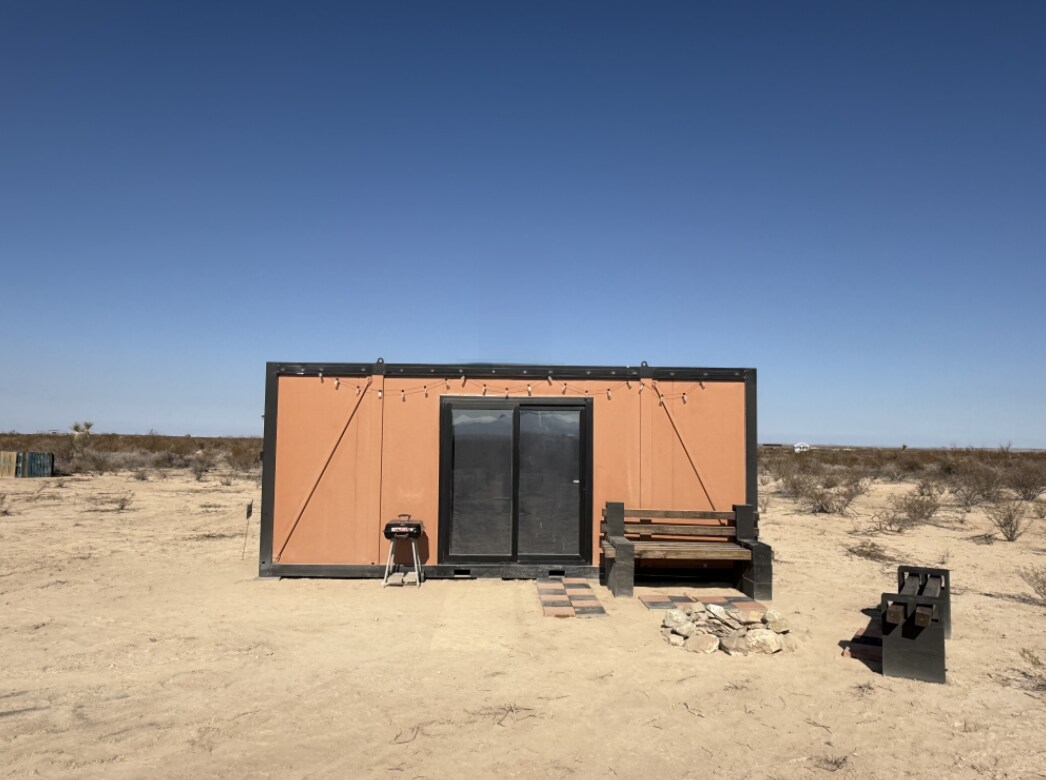 The exterior of a 20-foot insulated container home is displayed under a clear blue sky. A simple wooden bench and a propane grill are positioned in front, along with a small stone fire pit. The surrounding landscape is dry and sparse, typical of the desert environment.