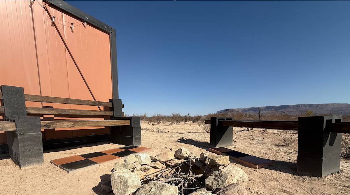 An outdoor seating area is positioned near a fire pit, made of rocks, providing an inviting space for gatherings. The surrounding landscape features arid terrain and distant mountains under a clear blue sky. Two rustic benches are arranged for comfort while enjoying the campfire.