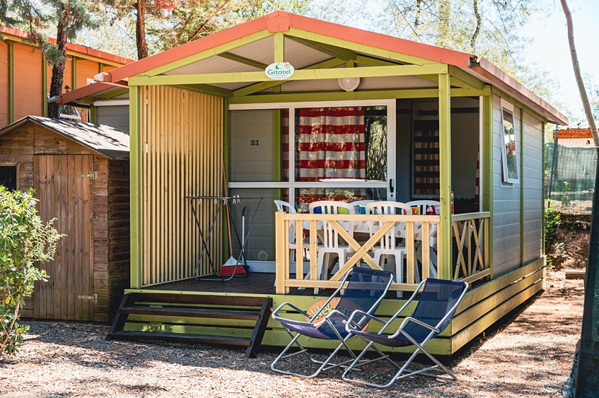 A bungalow is shown with a covered porch featuring a table and chairs for outdoor dining. Two lounge chairs are positioned on the porch, and a storage shed is visible to the left. Bright colors enhance the welcoming appearance of the structure.