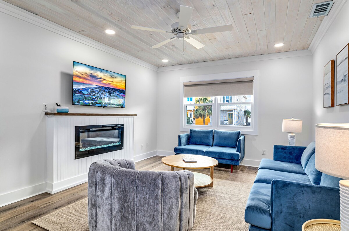 The living area features a ceiling fan and a flat-screen TV mounted on the wall. A light-colored area rug sits on the wood floor, accompanied by two blue sofas and a round wooden coffee table. An electric fireplace adds warmth to the space.