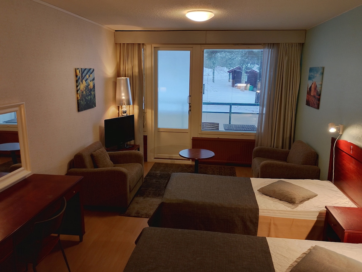 A functional room features two beds with grey blankets and a central pillow. Two armchairs and a small table are positioned near a window. A writing desk is located along one wall, complemented by a mirror. Light streams in, illuminating a peaceful view outside.
