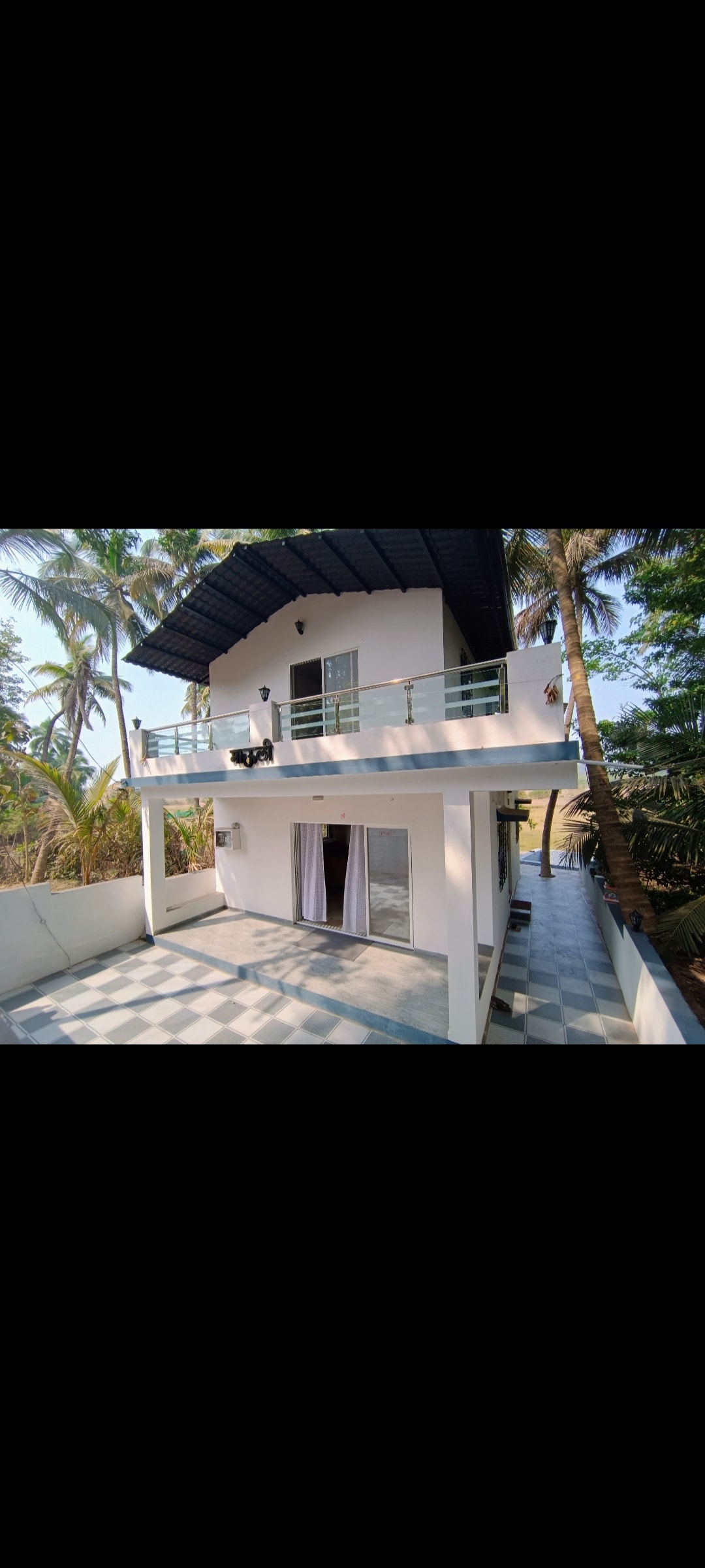 A modern two-story home is showcased with an open balcony that features glass railings. The ground level reveals a spacious terrace with tiled flooring, surrounded by tropical greenery, enhancing the outdoor living space.