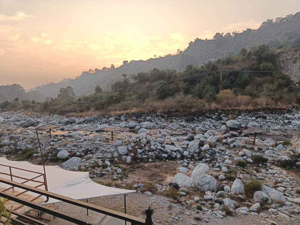 A serene landscape is captured during sunset, highlighting a rocky riverbed with smooth stones and sparse vegetation. Gentle hills rise in the background, with a soft glow from the setting sun illuminating the scene, creating a peaceful natural atmosphere.