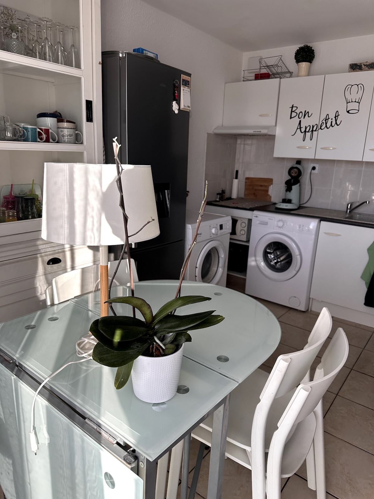 A modern kitchen area is displayed, featuring a clear glass dining table surrounded by white chairs. A potted orchid sits on the table beside a lamp. In the background, kitchen appliances include a refrigerator, washing machine, and dryer, with white cabinetry enhancing the space.