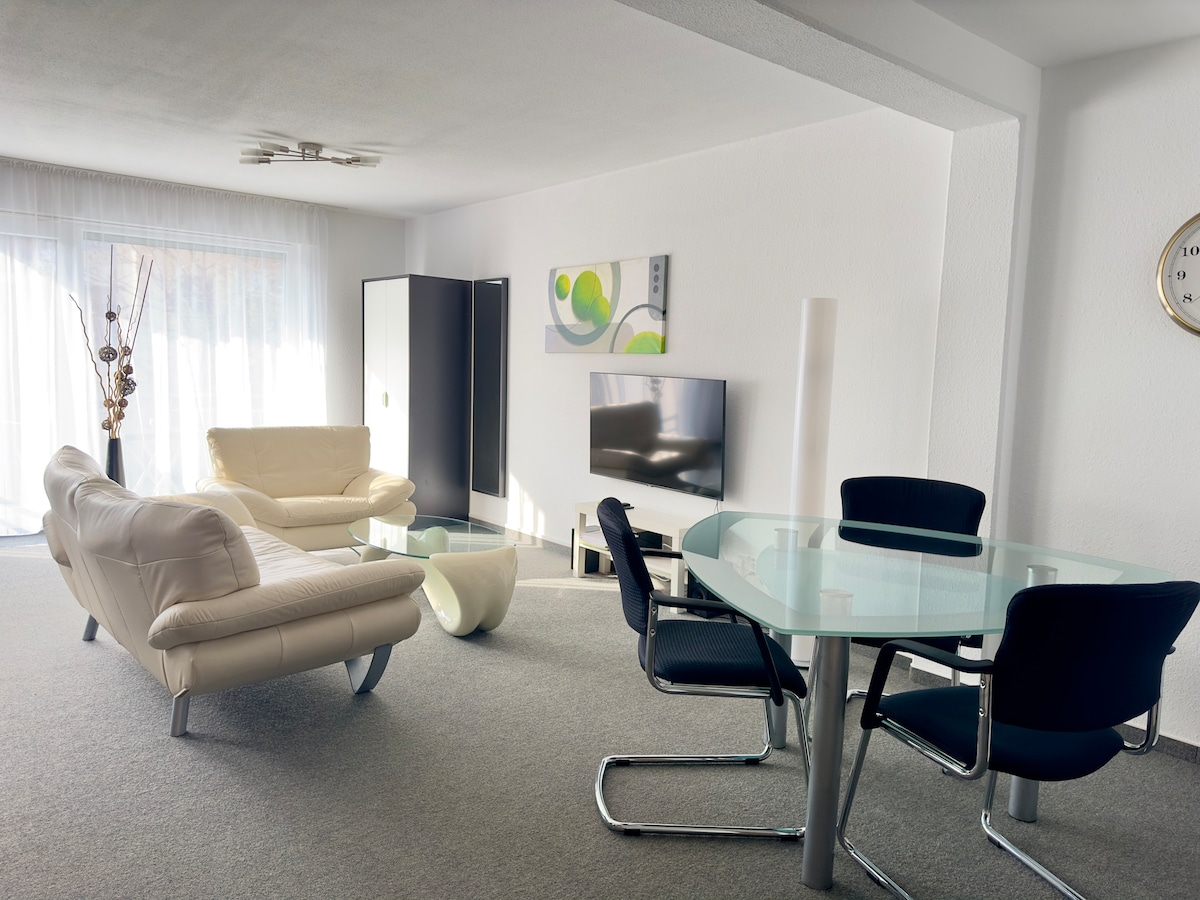 A modern living space is presented with sleek white sofas and a glass coffee table, offering a light and airy atmosphere. A dining area features a round glass table surrounded by black chairs. Natural light enters through large windows, enhancing the room's spaciousness.