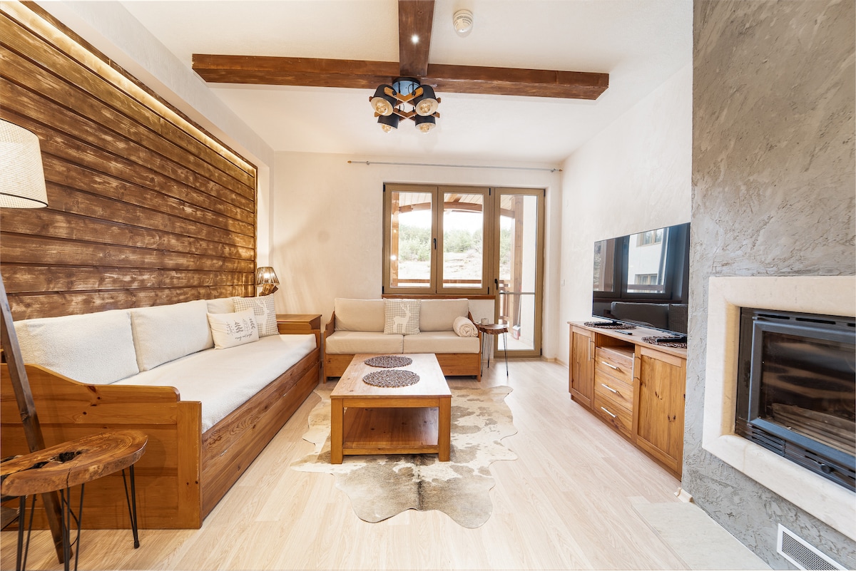 The spacious living room showcases a combination of wooden and neutral tones, featuring a comfortable L-shaped sofa and a wooden coffee table on a light area rug. Natural light streams through large windows, which provide access to the terrace. A fireplace and a flat-screen TV are also present.