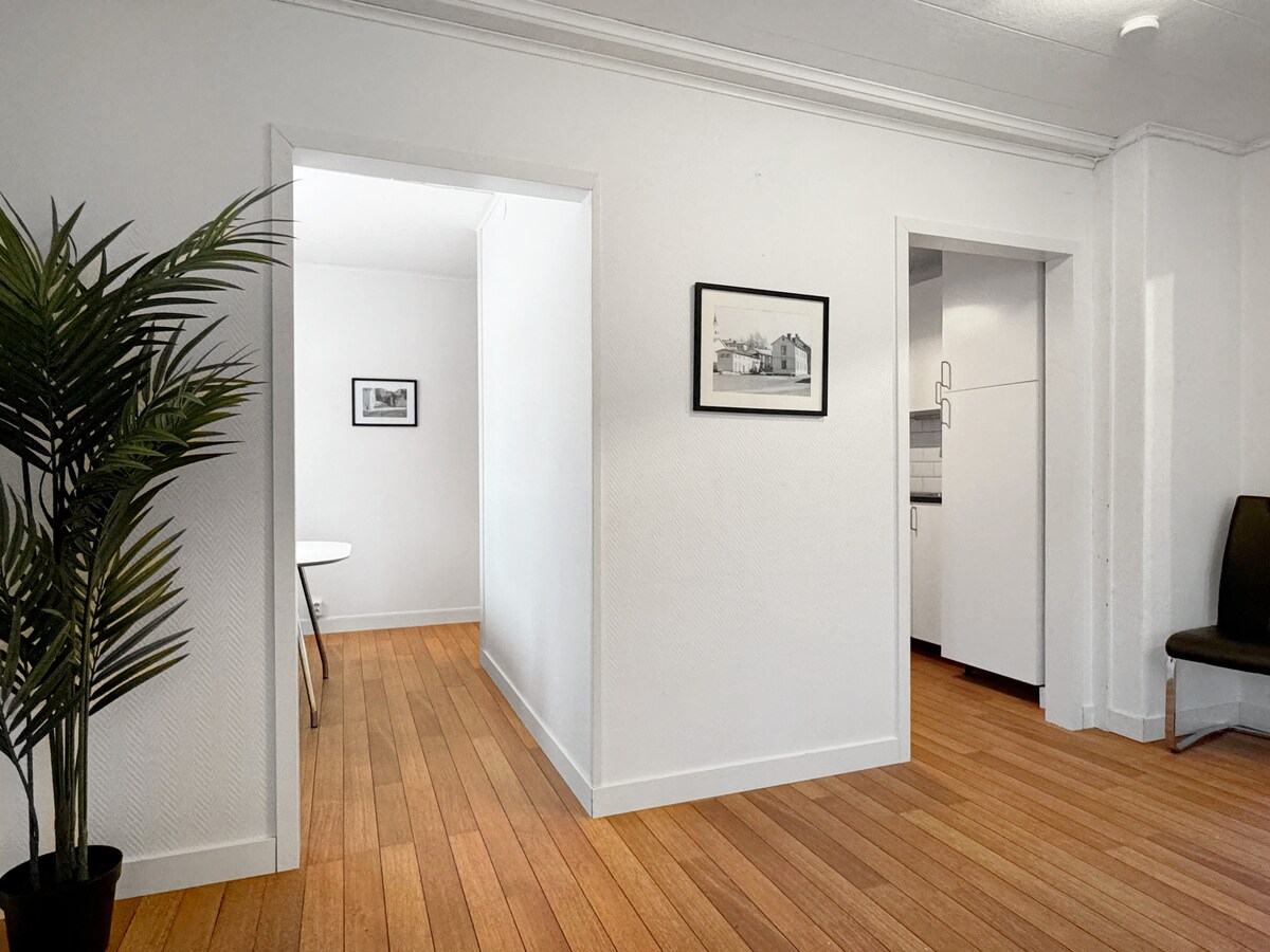 A bright hallway is visible, featuring smooth wooden flooring and white walls. Doorways lead to adjacent rooms. A small table with a light-colored surface is seen in the background, alongside a modern chair. Framed artwork is displayed on one wall.