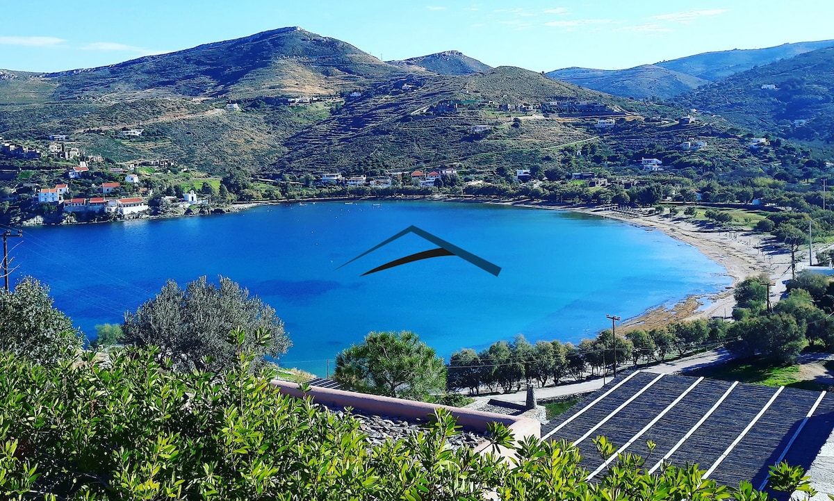 The image captures a serene view of Otzia beach, framed by lush greenery in the foreground. The calm blue waters gently reflect the surrounding hills and sky, creating a tranquil atmosphere. The beach features a gentle curve, leading to scattered homes along the coastline.