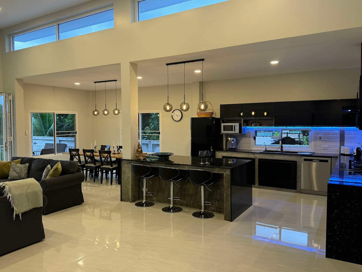 An open-plan living area features a modern kitchen with sleek black cabinets and stainless steel appliances. A spacious dining table is set nearby, illuminated by pendant lights. A comfortable seating area includes a dark sofa, with large windows allowing natural light to enter the space.