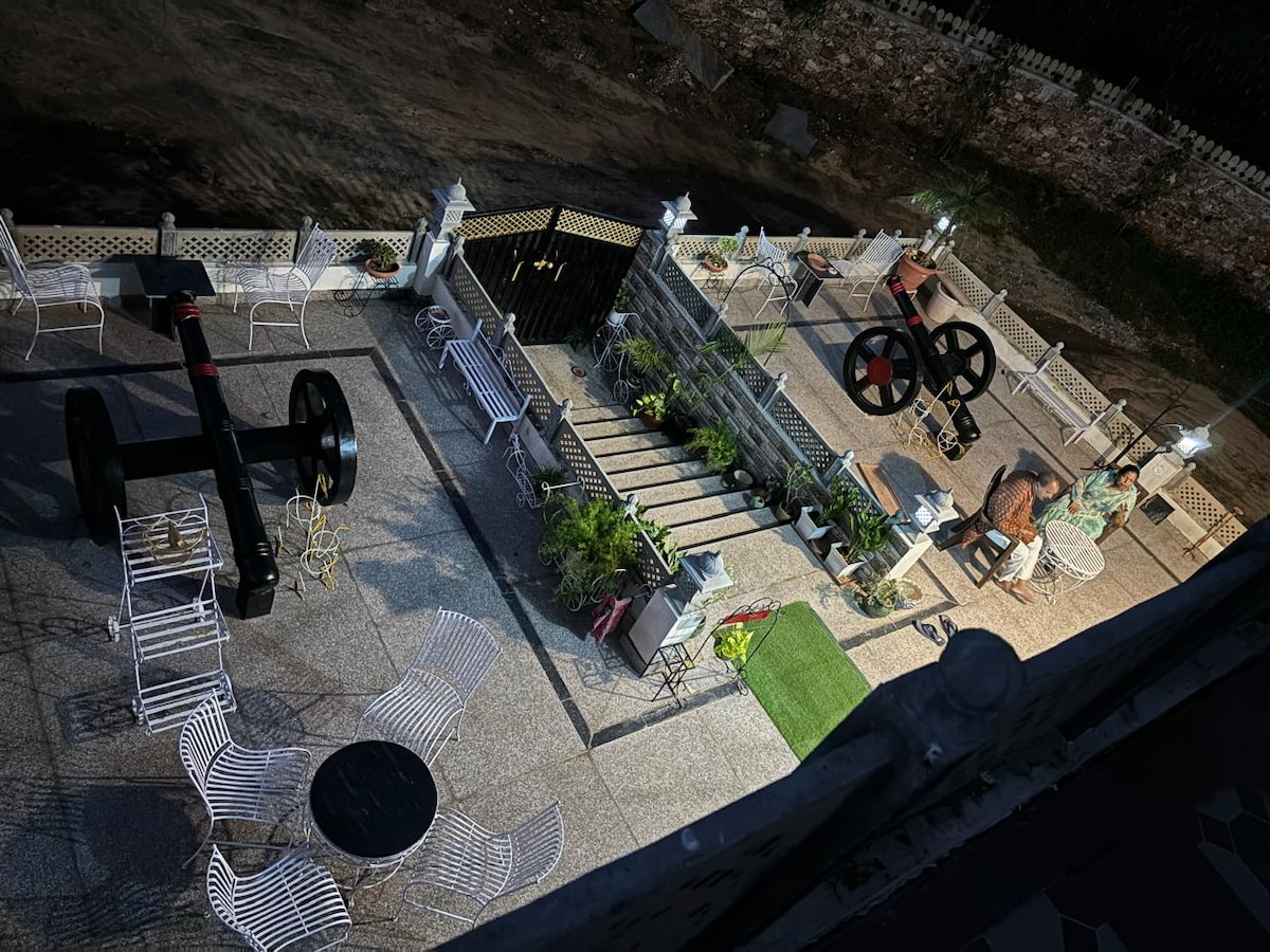 An outdoor courtyard is depicted, featuring white metal seating arranged around circular tables. Decorative elements include large black cannon replicas and strategically placed plants, enhancing the rustic ambiance. Soft lighting from nearby lamps illuminates the space, inviting relaxation in a serene environment.