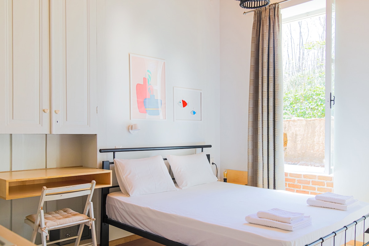 A bright bedroom features a spacious bed with white linens, accompanied by an open window with light curtains that allow natural light to enter. A simple desk and chair are placed against the wall, while colorful artwork adds a touch of creativity.
