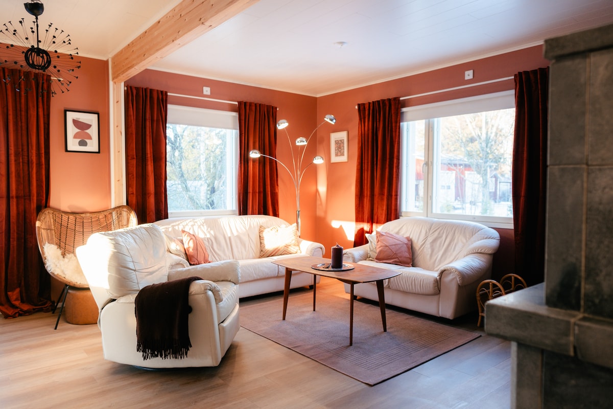 A well-lit living area features two white leather sofas arranged around a central table. Soft light fixtures illuminate the space, with large windows framed by rich red curtains offering views of the outdoors. A subtle area rug adds texture to the wooden floor.
