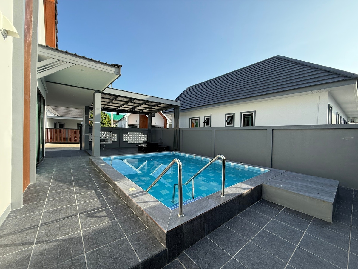 A private pool is surrounded by gray tiles, reflecting the clear sky above. Adjacent, a shaded area offers a roofed space for relaxation. The layout showcases a blend of outdoor and indoor living, enhancing the family-friendly atmosphere of the villa.