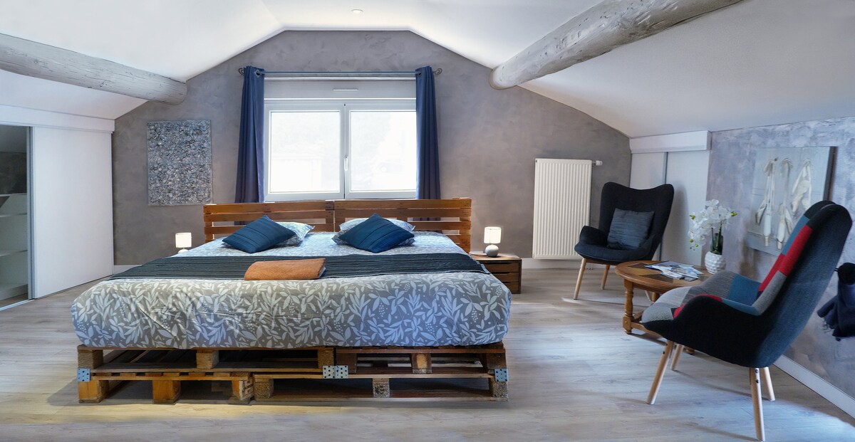 A spacious bedroom is featured, highlighted by a large bed with a patterned cover and decorative pillows. Two armchairs are positioned beside a bedside table, while a window provides natural light. The walls are finished in soft colors, and overhead beams contribute to a cozy ambiance.