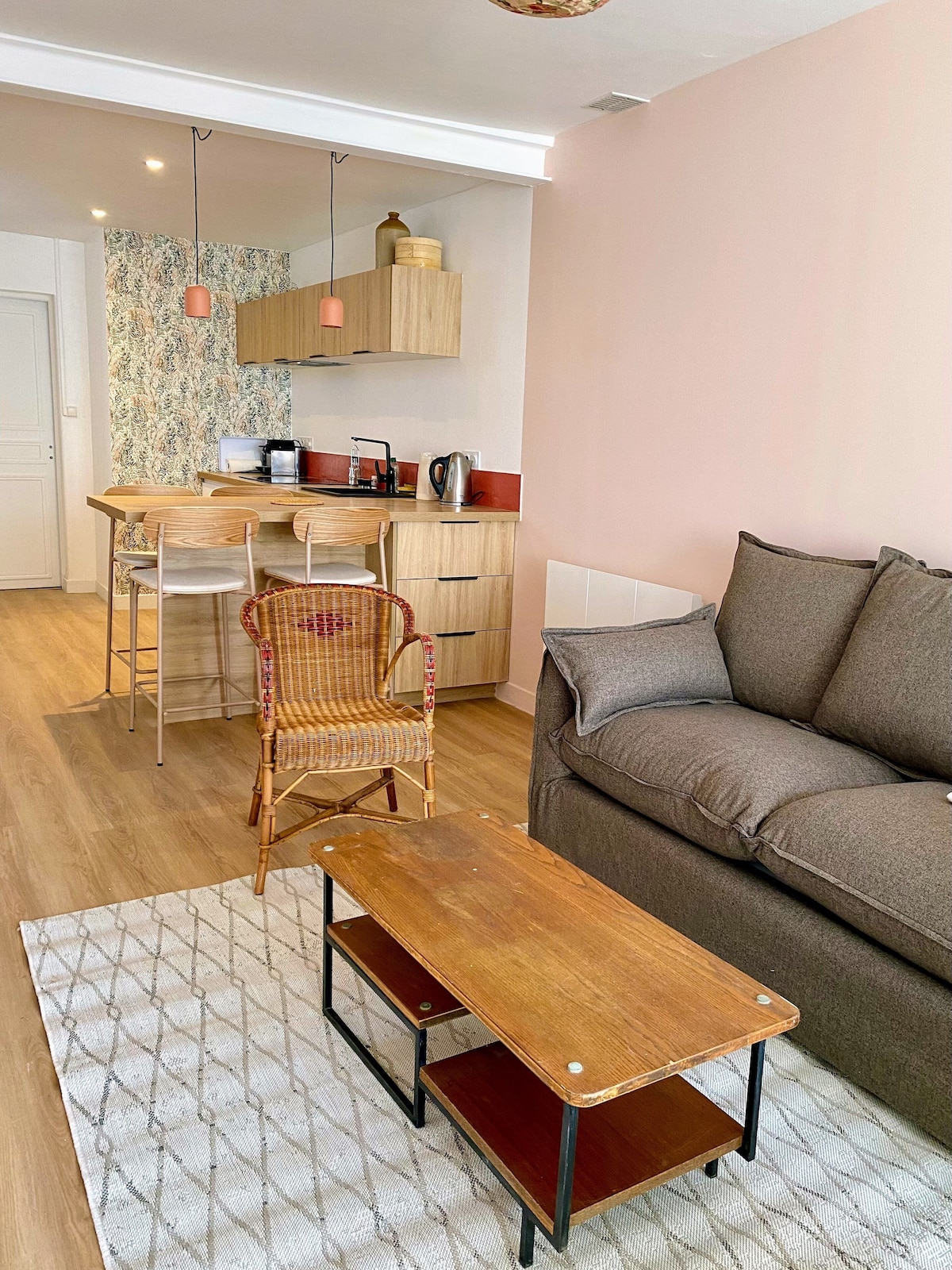 A cozy living area features a comfortable sofa and a wooden coffee table. A small dining area includes bar stools and modern kitchen appliances. The soft color palette and patterned wall add charm to the space, while natural light enhances the inviting ambiance.