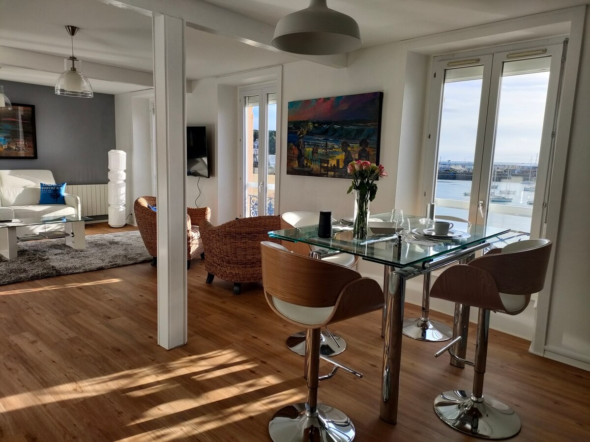 A modern dining area is furnished with a glass table and wooden chairs, complemented by a vase of flowers. Large windows allow natural light to fill the space, revealing views of the surroundings. A cozy living area is visible in the background, featuring comfortable seating.