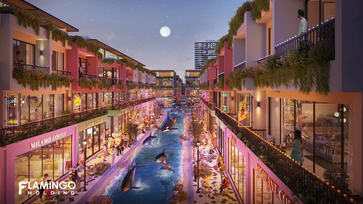 A vibrant commercial area is depicted, featuring colorful shopfronts and a waterway running through the center. Decorative lighting outlines the buildings and trees, contributing to a lively atmosphere. A few people can be seen on balconies and along the promenade, enjoying the surroundings.