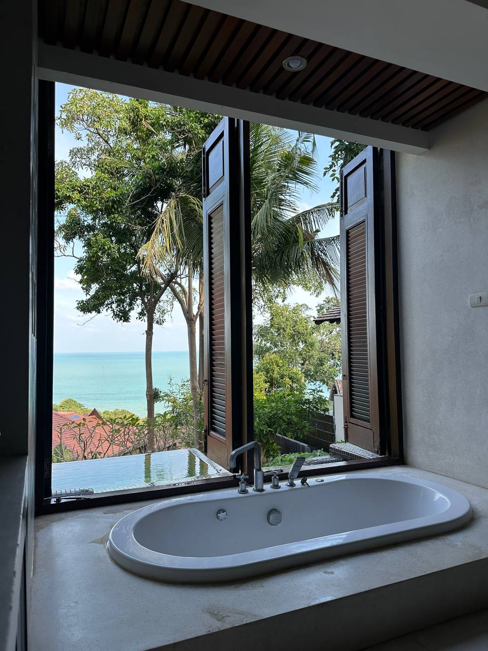 A modern bathtub is positioned next to large open windows, providing an unobstructed view of the sea and surrounding greenery. The natural light enhances the serene atmosphere, while elegant shutters add a touch of privacy.